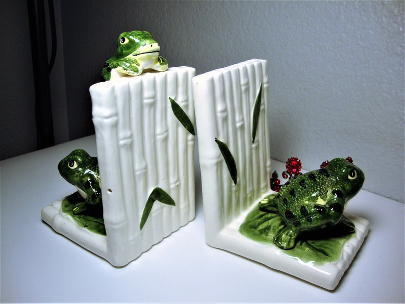 May include: A pair of ceramic bookends featuring green frogs on a white bamboo design. One frog is perched on top of the bamboo, while the other is sitting on a green leaf.