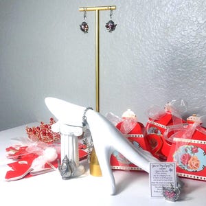May include: A display of tea-themed jewelry and gifts. Teapot earrings hang from a gold stand. A white hand-shaped jewelry holder displays a bracelet. Red teapot-shaped gift boxes and a crown are also present.