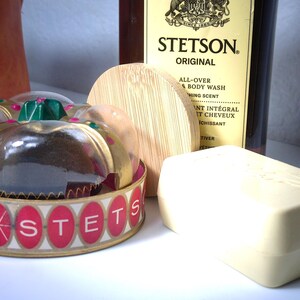 Pu&ograve; includere: Una collezione di prodotti per il bagno tra cui un contenitore di plastica trasparente con un'etichetta rossa e dorata che recita "SONSTETS", un coperchio di legno, una saponetta e una bottiglia di Stetson Original All-Over Body Wash.