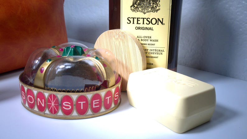 Pu&ograve; includere: Una collezione di prodotti per il bagno tra cui un contenitore di plastica trasparente con un'etichetta rossa e dorata che recita "SONSTETS", un coperchio di legno, una saponetta e una bottiglia di Stetson Original All-Over Body Wash.