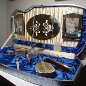 May include: A vintage vanity set in a blue and white case with a mirror, brushes, and a bottle of perfume. The case is lined with blue satin and has a gold trim. The case is open and the contents are visible.