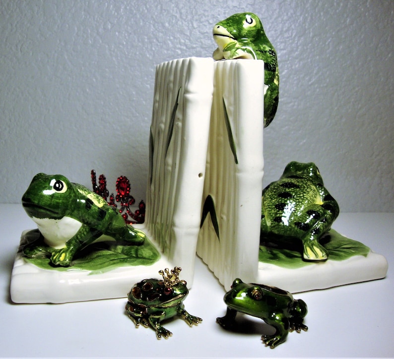 May include: A set of ceramic frog bookends with green and white painted details. The bookends are shaped like frogs sitting on a green base with bamboo stalks. Two smaller jeweled frog figurines are also included.