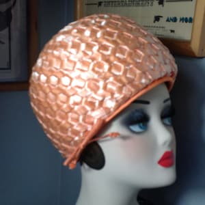 May include: A peach-colored vintage swim cap with a geometric pattern. The cap has a rounded shape and a contrasting orange trim. The cap is displayed on a mannequin head with red lipstick and blue eyes.