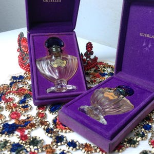 May include: Two vintage Guerlain perfume bottles in purple velvet boxes. The clear glass bottles have a unique shape and are topped with dark blue stoppers. The boxes are open, revealing the perfume bottles inside. The Guerlain logo is visible on the box.