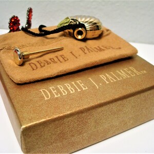 May include: A gold-colored perfume bottle in the shape of a seashell with a black cord and a gold-colored stopper. The bottle is in a brown suede pouch with the text "Debbie J. Palmer" printed on it. The pouch is sitting on top of a gold-colored box with the text "Debbie J. Palmer" printed on it.