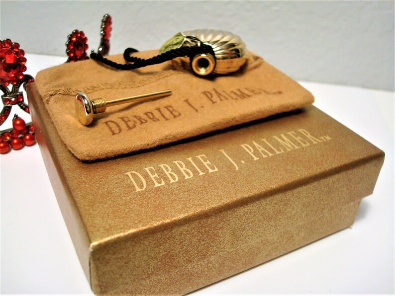 May include: A gold-colored perfume bottle in the shape of a seashell with a black cord and a gold-colored stopper. The bottle is in a brown suede pouch with the text "Debbie J. Palmer" printed on it. The pouch is sitting on top of a gold-colored box with the text "Debbie J. Palmer" printed on it.