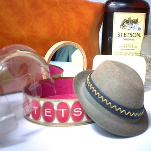 Pu&ograve; includere: Una collezione di prodotti e accessori per la cura della persona vintage. Include un cappello Stetson marrone con fascia blu navy e oro, una bottiglia di dopobarba Stetson Original, un flacone di profumo e una pochette in pelle.