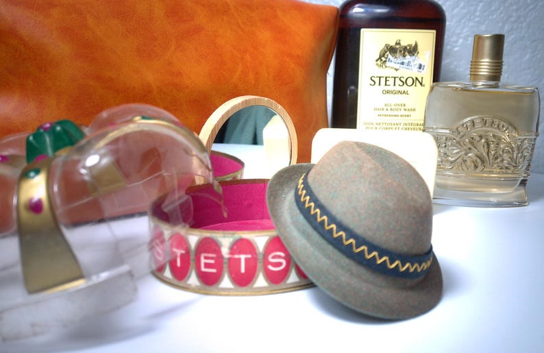 Pu&ograve; includere: Una collezione di prodotti e accessori per la cura della persona vintage. Include un cappello Stetson marrone con fascia blu navy e oro, una bottiglia di dopobarba Stetson Original, un flacone di profumo e una pochette in pelle.