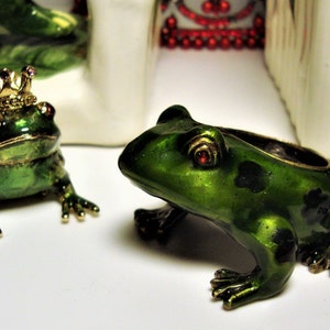 May include: Two green frog figurines with gold accents. One frog is wearing a gold crown with pink jewels. Both frogs have red eyes.