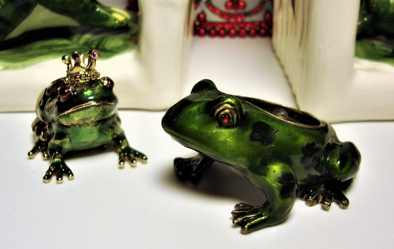 May include: Two green frog figurines with gold accents. One frog is wearing a gold crown with pink jewels. Both frogs have red eyes.