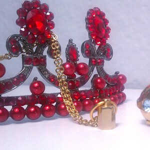 May include: A decorative crown with red jewels and a gold chain, next to a gold perfume bottle with blue and clear gems. The crown has a dark metal frame and is adorned with red jewels.