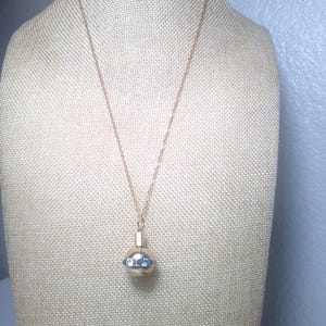 May include: A gold-toned necklace with a pendant. The pendant is a small, round, gold-colored container with two blue gemstones and two clear gemstones. The necklace is displayed on a beige mannequin.