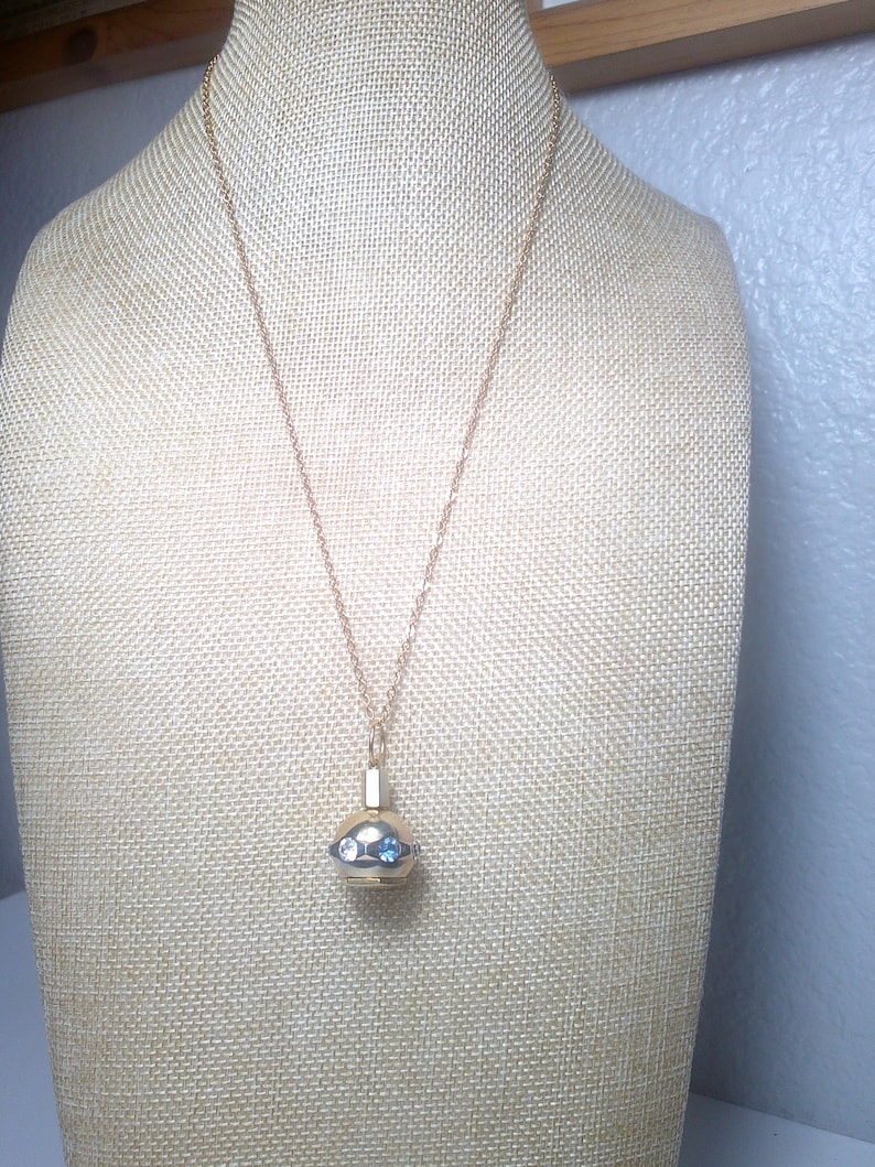 May include: A gold-toned necklace with a pendant. The pendant is a small, round, gold-colored container with two blue gemstones and two clear gemstones. The necklace is displayed on a beige mannequin.