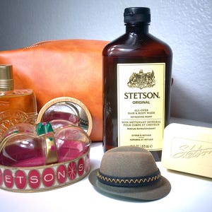 Pu&ograve; includere: Un assortimento di prodotti per la cura della persona, tra cui una bottiglia di Stetson Original Hair & Body Wash, una saponetta, un cappello piccolo e un flacone di profumo. Sono presenti anche un astuccio in pelle e una corona decorativa. I prodotti sono disposti su una superficie bianca.