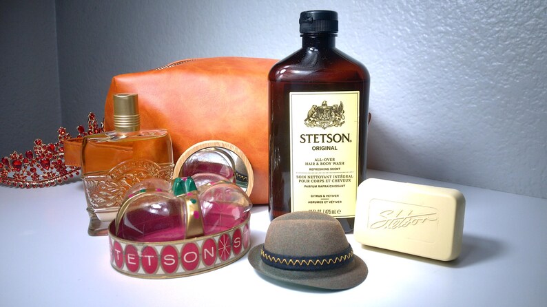 Pu&ograve; includere: Un assortimento di prodotti per la cura della persona, tra cui una bottiglia di Stetson Original Hair & Body Wash, una saponetta, un cappello piccolo e un flacone di profumo. Sono presenti anche un astuccio in pelle e una corona decorativa. I prodotti sono disposti su una superficie bianca.