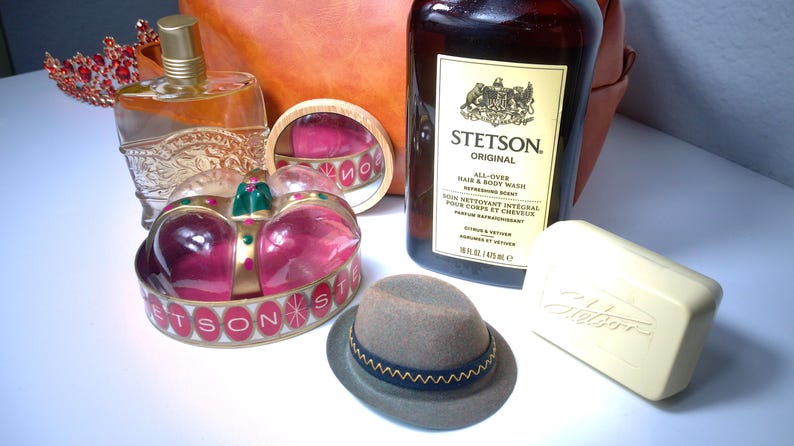 Pu&ograve; includere: Un assortimento di articoli per la cura della persona e decorativi. Include una bottiglia di bagnoschiuma e shampoo Stetson Original, un piccolo cappello fedora grigio con una fascia blu, una saponetta, un flacone di profumo e un portagioie a forma di corona.