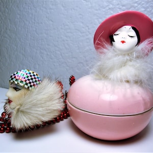 May include: Two vintage ceramic figurines of women with closed eyes. The figurine on the left is wearing a colorful checkered hat and a white fur collar. The figurine on the right is wearing a pink hat and a white feather boa. Both figurines are sitting on a pink ceramic base.