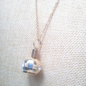 May include: A gold-colored pendant necklace featuring a small, round perfume bottle design. The bottle is adorned with blue and clear crystals. The necklace has a delicate chain and a simple clasp.