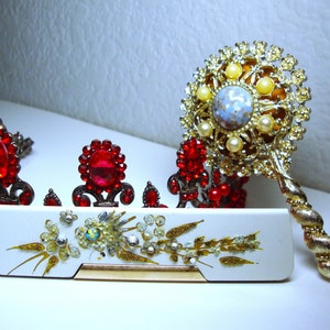May include: A set of two vintage hair combs with red and gold accents. The combs feature large red jewels and intricate gold designs. The combs are displayed on a white surface with a gold and white design.