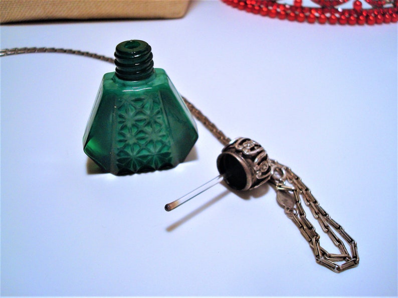 May include: A green glass bottle with a starburst pattern and a silver chain attached to a silver stopper. The bottle is empty and the stopper is removed.