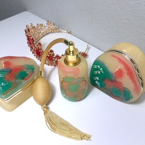 May include: A collection of vintage vanity items, including a heart-shaped box, a perfume bottle with a tassel, a small crown, and a compact. The items feature a cream and gold color scheme with painted floral designs in red, green, and peach.