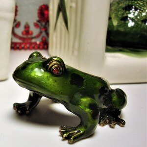 May include: A small green frog figurine with red eyes and gold accents. The frog is sitting on a white surface.