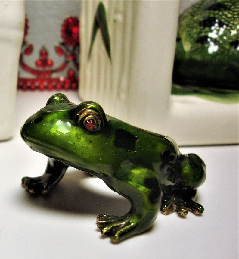 May include: A small green frog figurine with red eyes and gold accents. The frog is sitting on a white surface.