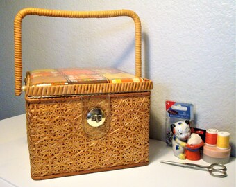 May include: A rectangular sewing basket with a woven wicker body, a tall arched handle, and a hinged lid featuring a plaid fabric top in shades of orange, brown, and yellow. The basket includes a silver-tone metal latch and a small pin cushion.