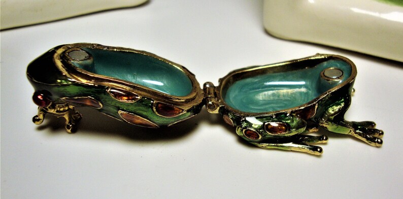 May include: A green frog-shaped trinket box with gold accents and red and orange spots. The box is open, revealing a turquoise interior.
