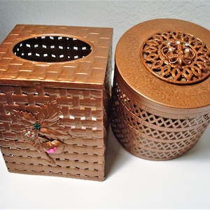 May include: Two copper-colored tissue box holders. One is square with a woven design and a floral embellishment. The other is round with a decorative lid and a cut-out design.