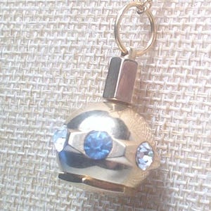 May include: A gold-colored pendant necklace featuring a round, orb-shaped charm. The charm is adorned with three blue and white gemstones. The pendant hangs from a delicate gold chain, creating an elegant accessory.
