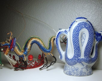 May include: A blue and white porcelain teapot shaped like a dragon. The teapot has a spout and handle. A blue and gold wire dragon with red and white beads is also in the image.