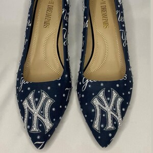 May include: A pair of navy blue flats with white and silver embellishments. The shoes feature the New York Yankees logo and the words "Dream Pairs" on the insole.