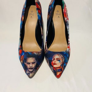 Può includere: Un paio di scarpe nere con tacco alto e punta a punta. Le scarpe sono ricoperte da una stampa colorata con i personaggi Joker e Harley Quinn del film Suicide Squad. La stampa include i colori rosso, blu, verde e giallo. Le scarpe hanno un tacco nero e una suola nera. L'interno delle scarpe ha un'etichetta che dice "Call It Spring."