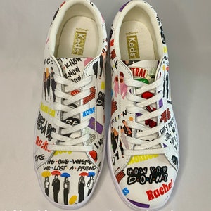 May include: A pair of white sneakers with a colorful print featuring characters and quotes from the TV show "Friends". The sneakers have a gold "Keds" logo on the tongue.