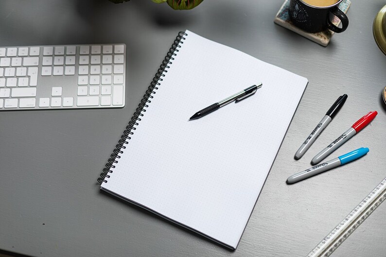May include: A white spiral bound notebook with a black pen and three highlighters on a gray surface. The highlighters are black, red, and blue.