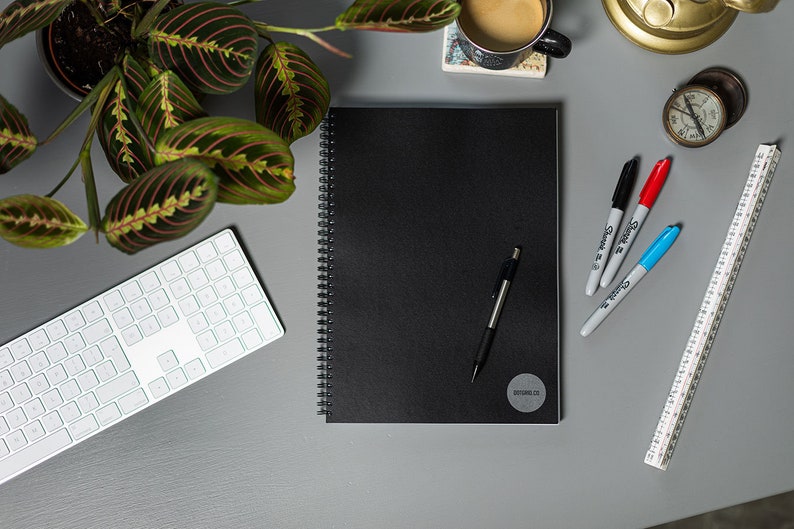 May include: A black spiral bound notebook with a silver pen and a white sticker with the text "BEYONDCO" on the cover. The notebook is on a gray surface with a white keyboard, a cup of coffee, a ruler, and three markers.
