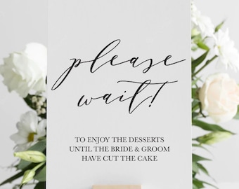May include: White card with black calligraphy that reads "please wait!" and the text "To enjoy the desserts until the bride & groom have cut the cake". The card is propped up on a small wooden block.