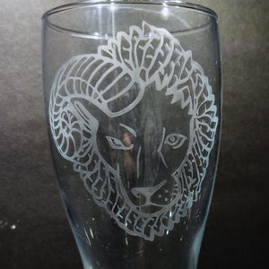 Pode incluir: Uma caneca de cerveja de vidro transparente com um desenho gravado de uma cabeça de leão com juba.