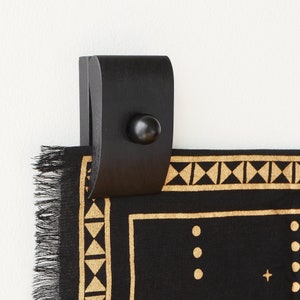 May include: Black wooden wall hanging with a black and gold geometric pattern. The hanging has a black wooden knob and a fringe border.