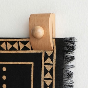 May include: A wooden wall hook with a rounded knob holds a black textile with a gold geometric border. The textile has a fringed edge. The hook is attached to a white wall.