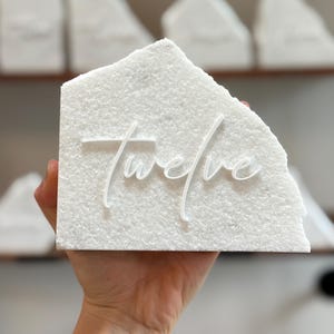 May include: A white textured block with the word "twelve" in a cursive font. The block has a rough, uneven edge. Other similar blocks are visible in the background, suggesting a set of table numbers or decorative pieces.