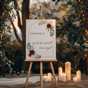 Könnte beinhalten: Ein weißes Schild mit schwarzem Rahmen und einem Bogen-Design mit roten und weißen Rosen und grünen Blättern. Das Schild lautet "Veronica, quieres casarte conmigo?"  Das Schild steht auf einem Holzstaffelei vor einem verschwommenen Hintergrund aus Grün und Lichtern.
