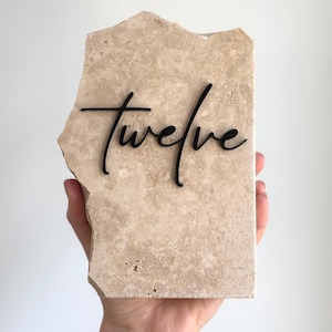May include: A rectangular piece of beige travertine stone with rough, uneven edges. The word twelve is written across the center in black, lowercase, cursive script lettering. The stone has a natural, porous texture with varied light brown tones.