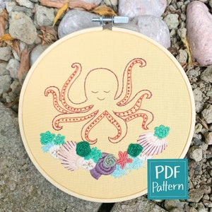 May include: Embroidered hoop art featuring a red octopus with closed eyes and a smiling mouth surrounded by a wreath of green, pink, and purple flowers and seashells. The hoop is yellow and has a wooden frame. The text "PDF Pattern" is visible in the lower right corner.