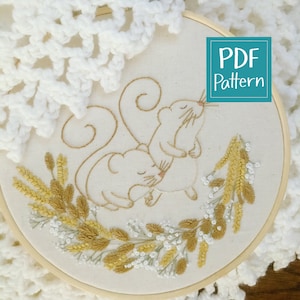 May include: Embroidered hoop art featuring two white mice with curled tails, nestled amongst a wreath of golden wheat and white flowers. The hoop is made of wood and has a white fabric background. The text "PDF Pattern" is displayed in a teal box.