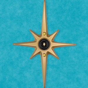 May include: A gold-colored starburst-shaped door knocker with a black circular button in the center. The knocker is mounted on a blue wall.