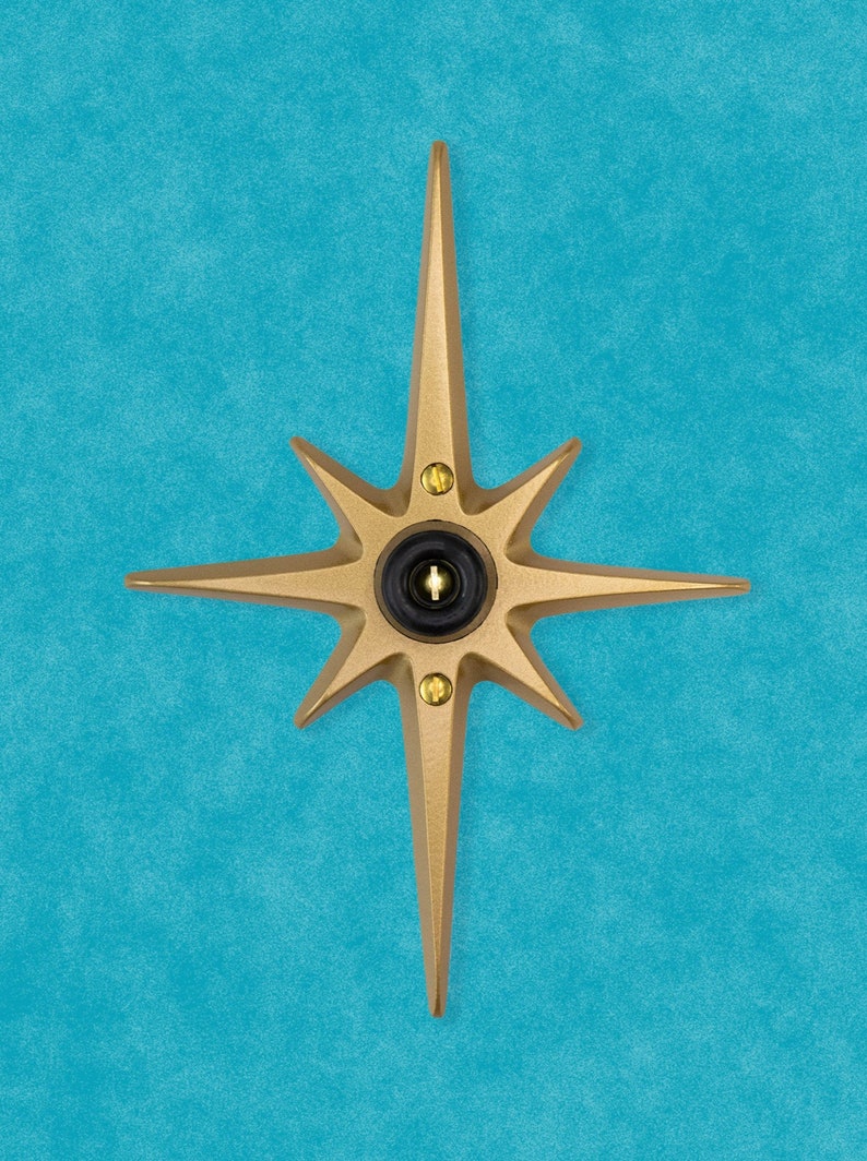 May include: A gold-colored starburst-shaped door knocker with a black circular button in the center. The knocker is mounted on a blue wall.