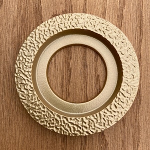 May include: A gold-colored, circular decorative object with a textured outer ring and a smooth inner ring. The object is set against a wooden background, creating a contrast in textures and colors. The design is simple yet elegant.