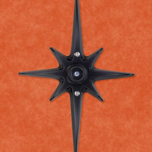 May include: A black metal starburst with eight points, each point ending in a sharp tip. The starburst has a circular center with a small hole in the middle.